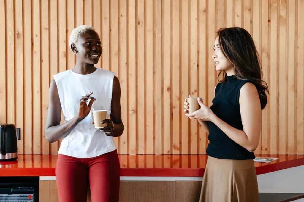 Happy multiracial women with cups of coffee smiling and discussing job in contemporary office